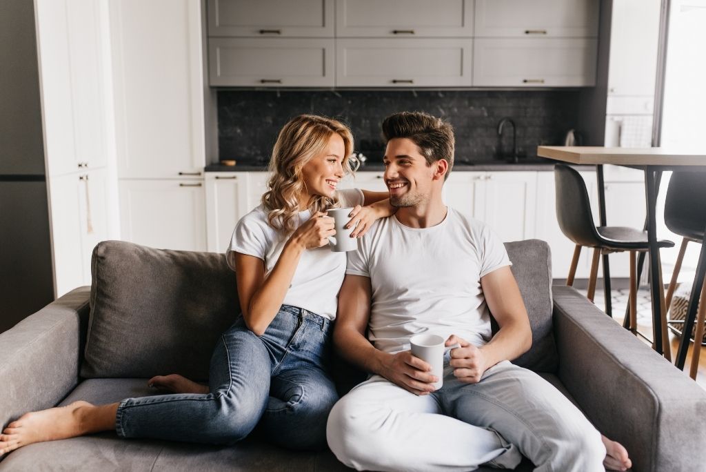 pareja de enamorados sentados en el sofá sosteniendo tazas mirando el uno al otro y sonriendo. pareja romántica disfruta de la mañana juntos en casa